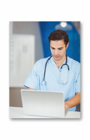 Professional wearing scrubs and stethoscope typing on silver laptop on white desk in nurse station