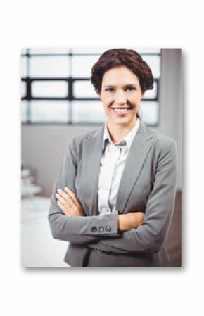 Businesswoman wearing grey suit posing crossing arms at desk with monitor and grid window in office