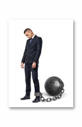 A sad businessman looks down while shackled to a large iron ball with a chain to his ankle.