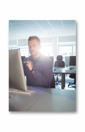 Office worker raising eyeglasses facing large monitor with keyboard in sunlit office rows of desks