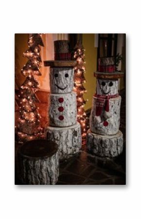 Snowmen constructed from tree logs, wearing hats, standing next to illuminated Christmas trees
