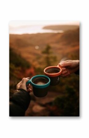 Vertical of two people holding mugs with hot coffee on a blurry background of autumn landscape