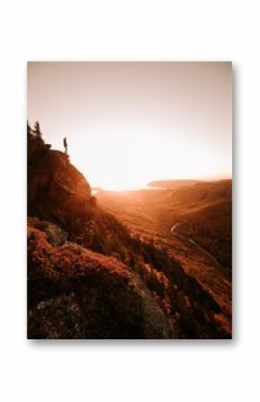 Vertical shot of a person standing on a cliff, above a river in mountains in autumn at sunset