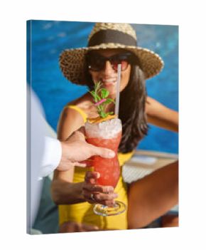 Cheerful young female tourist in stylish straw hat and sunglasses receiving fresh cold fruit beverage from unrecognizable barmen while enjoying summer vacation at poolside in resort