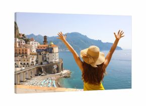 Summer holiday in Italy. Back view of young woman with straw hat and yellow dress with raised arms looking at Atrani village, Amalfi Coast, Italy
