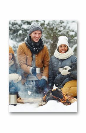 Joyful young people in winterwear sitting in snow arounf fire and having hot drinks