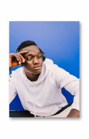 Young afro American black man wearing a white sweatshirt and headphones seated on a blue wall looking to the side. Modern look