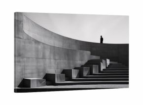 A black and white photograph of a modern, minimalist architectural structure with curved and stepped concrete elements. A solitary figure stands at the top of the structure.