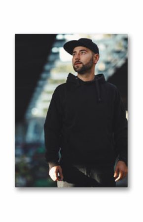 Portrait of a young bearded man with a black hat ain black hoodie in the city