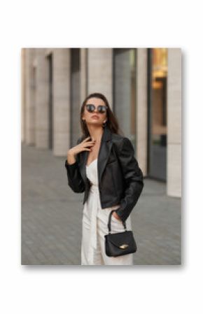 Young stylish woman in white overall, sneakers, black leather jacket and sunglasses holding small handbag and standing at city street. Fashion style outdoor portrait