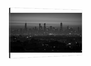 Moody black and white panorama of a major city skyline with illuminated skyscrapers seen through atmospheric haze at night.