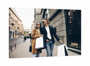 Fashionable couple walking in city with shopping bags, laughing and enjoying time together. Man in black jacket and white shirt,  Asian woman in camel coat and sunglasses. Black friday sales.