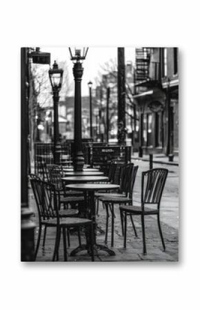 A black and white photograph of an empty outdoor café with vintage street lamps, evoking a sense of quiet urban solitude on a serene city street corner.