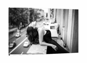 Black white portrait of young woman in elegant silk black dress lookingto a city from a balcony, fashion beauty photo