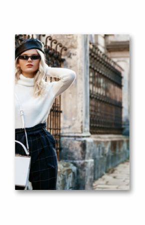 Outdoor street fashion portrait of elegant lady wearing stylish autumn outfit: black leather beret, sunglasses, white turtleneck, checkered high waist trousers, small bag, posing in city. Copy space