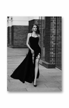  High fashion. Sexy girl in classic evening dress. Black and white photo. Luxury vintage woman with long, bare leg. Beautiful retro Lady. Glamour queen. Noir style chic model walks on old city street 