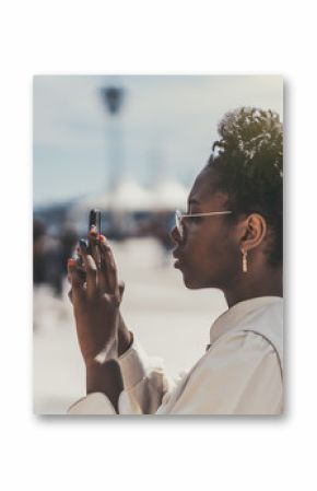 Side view of a cute black hipster girl in spectacles and white trench taking pics of the camera of her smartphone  charming young African female tourist is shooting attractions on her cellphone