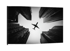 A black and white perspective of a plane flying above towering city buildings.  A low angle view looks up at the aircraft amidst skyscrapers