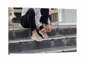 Woman sitting on outdoor stone steps tying shoelaces on pale pink athletic shoes with smartwatch