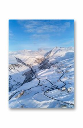 Aerial of snowy mountain range on winter sunrise at ski resort. Drone above mountains valley and village with curvy road at sunset. Caucasus peaks skyline in the pink twilight afterglow.