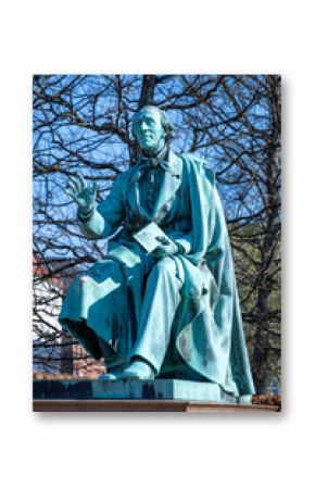 A detailed bronze statue of Hans Christian Andersen sits in the serene Rosenborg Castle Gardens, Copenhagen, surrounded by trees and a clear sky, representing the famous storyteller's legacy.