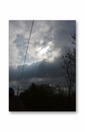 A dramatic sky with bright light breaking through dark, ominous clouds, silhouetting power lines and bare trees in the foreground.