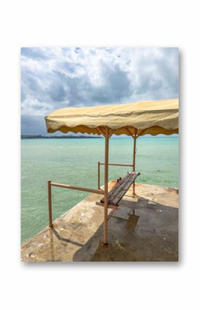 Rusty wooden bench with canopy on concrete pier by metal fence overlooking sea, in cloudy weather, perfect for themes of isolation