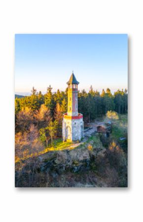 The Stepanka Lookout Tower stands tall against a clear spring sky in Korenov, surrounded by lush greenery. Sunlight illuminates the tower, highlighting its architectural beauty.