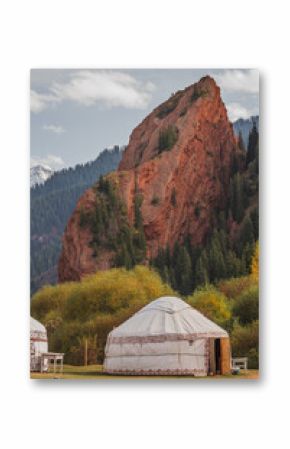 Traditional yurts stand in a picturesque valley, framed by towering red rock formations and lush evergreens. The stunning autumnal foliage adds a vibrant touch to this breathtaking scene in