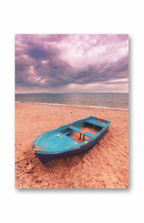 Abandoned blue fishing boat on sandy beach under dramatic cloudy sky. Weathered wooden vessel near calm sea, moody atmosphere with pink sunset tones and distant horizon.