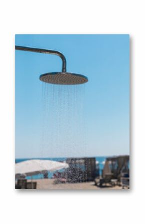 A metal shower head dispenses water in a beach setting with a bright blue sky above. Sunbeds and umbrellas are visible near the shore, inviting relaxation and refreshment