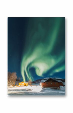 Northern lights light up the entire sky under small wooden cottages with fire inside. Cozy and fantasy evening in Northern Norway