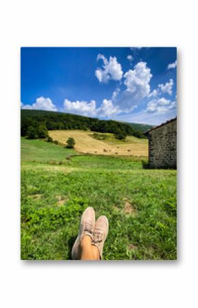 Resting bare feet on lush green grass, overlooking hay bales, stone farmhouse, wooded mountain beneath cloudy blue sky