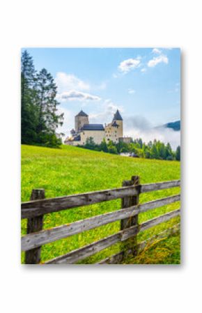 Mauterndorf Castle stands majestically against a backdrop of clear blue skies and rolling hills, surrounded by vibrant green fields and tall trees, creating a picturesque Austrian scene.