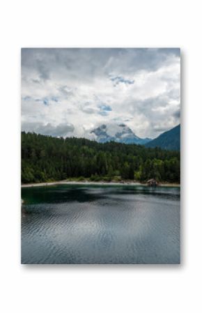Scenic view of Blindsee lake in Tyrol, Austria, a popular diving paradise. Clear alpine water with ripples in the foreground, surrounded by dense green forest and dramatic mountain peaks in clouds.