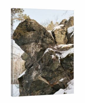 Large rugged rock formation covered with patches of snow standing in winter forest, bare trees and evergreen branches surrounding rocky cliff, natural outdoor landscape scene