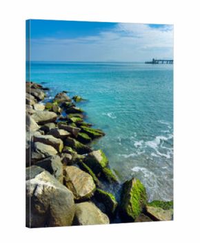 Vibrant coastal scene showcasing rocky shoreline and tranquil waters of the seaside at midday with a distant pier under a bright blue sky