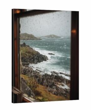 View through a rain-speckled window reveals rocky Irish coastline and turbulent sea under grey light. The scene feels windswept, moody, and deeply atmospheric.