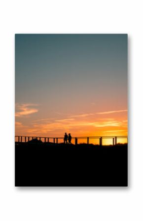  Silhouetted couple walks along a boardwalk at sunset, framed by vivid orange and blue skies. A serene, atmospheric moment with ample copy space for text overlay or creative design