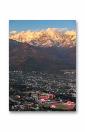 A captivating aerial shot of Dharamshala city, with the snow-covered Dhauladhar mountains glowing in the evening light and the beautiful Dharamshala Cricket Stadium shining in the city’s center.