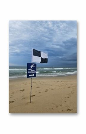 Cloudy Beach Day: black and white flag marks designated surfcraft area, waves gently crashing as footprints scatter across the beach