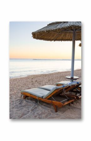 Relaxing beachside loungers under a straw umbrella at sunset by the calm ocean waves
