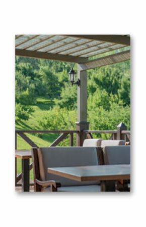A cozy terrace with soft chairs and wooden furniture, a canopy and lantern, set against a green hill and dense forest lit by bright sunlight.