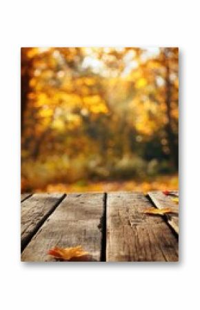 Rustic wooden table with scattered autumn leaves against blurred background of sunlit fall forest. Perfect for seasonal product display or cozy autumn theme
