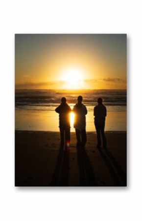 Three teenage girls silhouetted quietly on the dark sands of Muriwai Beach, gazing at a golden sunset as it reflects on the ocean waves and clouds in a serene evening atmosphere