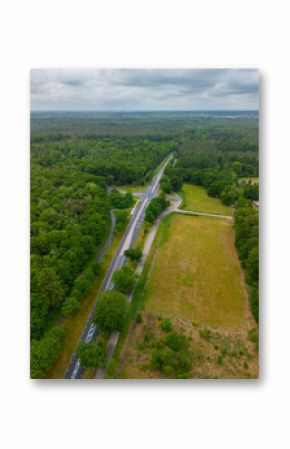 A drone view of a road cutting through dense green forest, with open grassy fields on one side and residential houses tucked among the trees, capturing a peaceful blend of nature and infrastructure.