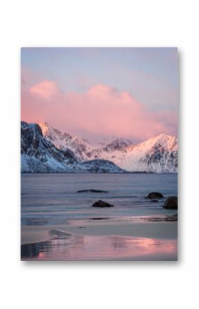 Colorful winter sunrise at Haukland Beach, Lofoten Islands, Norway. Snowy mountain and pink clouds reflect in the calm sea and wet sand, creating a peaceful Arctic landscape.