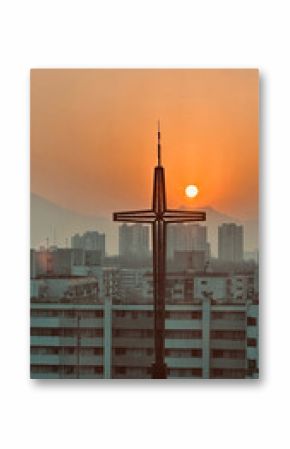 Metal cross structure silhouetted by a rising sun over a cityscape with high-rise buildings and distant mountains.