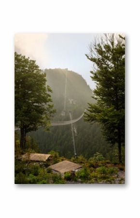 Suspension footbridge Highline179 in Tirol, Austria, spanning across a deep forest valley with misty mountains and dramatic sunlight breaking through the clouds.