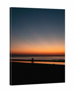 Silhouette of cyclist riding along beach at sunset with vibrant orange sky reflecting on calm ocean water